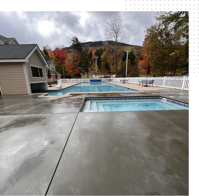 Outdoor commercial swimming pool and integrated square hot tub with concrete deck, surrounded by fall foliage and a mountain resort backdrop