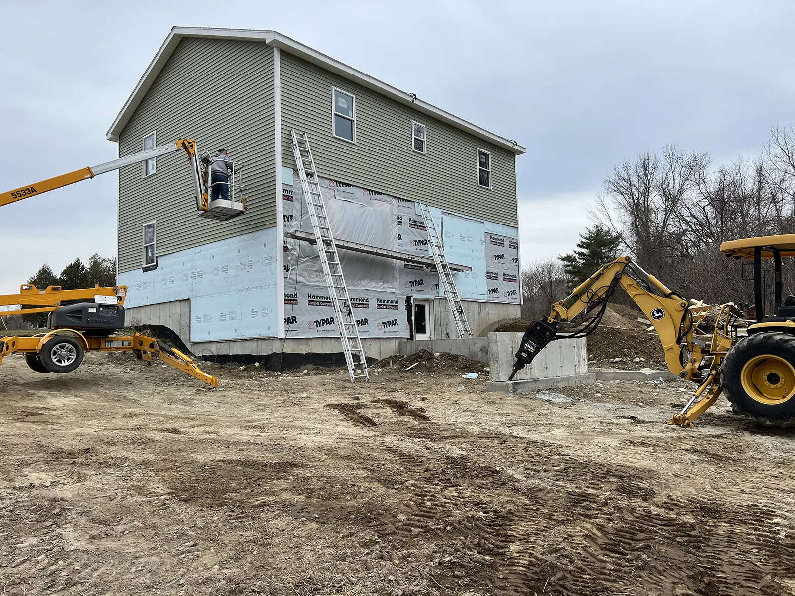 Crew on a boom lift installing windows on a two-story home, with ladders and a backhoe on site