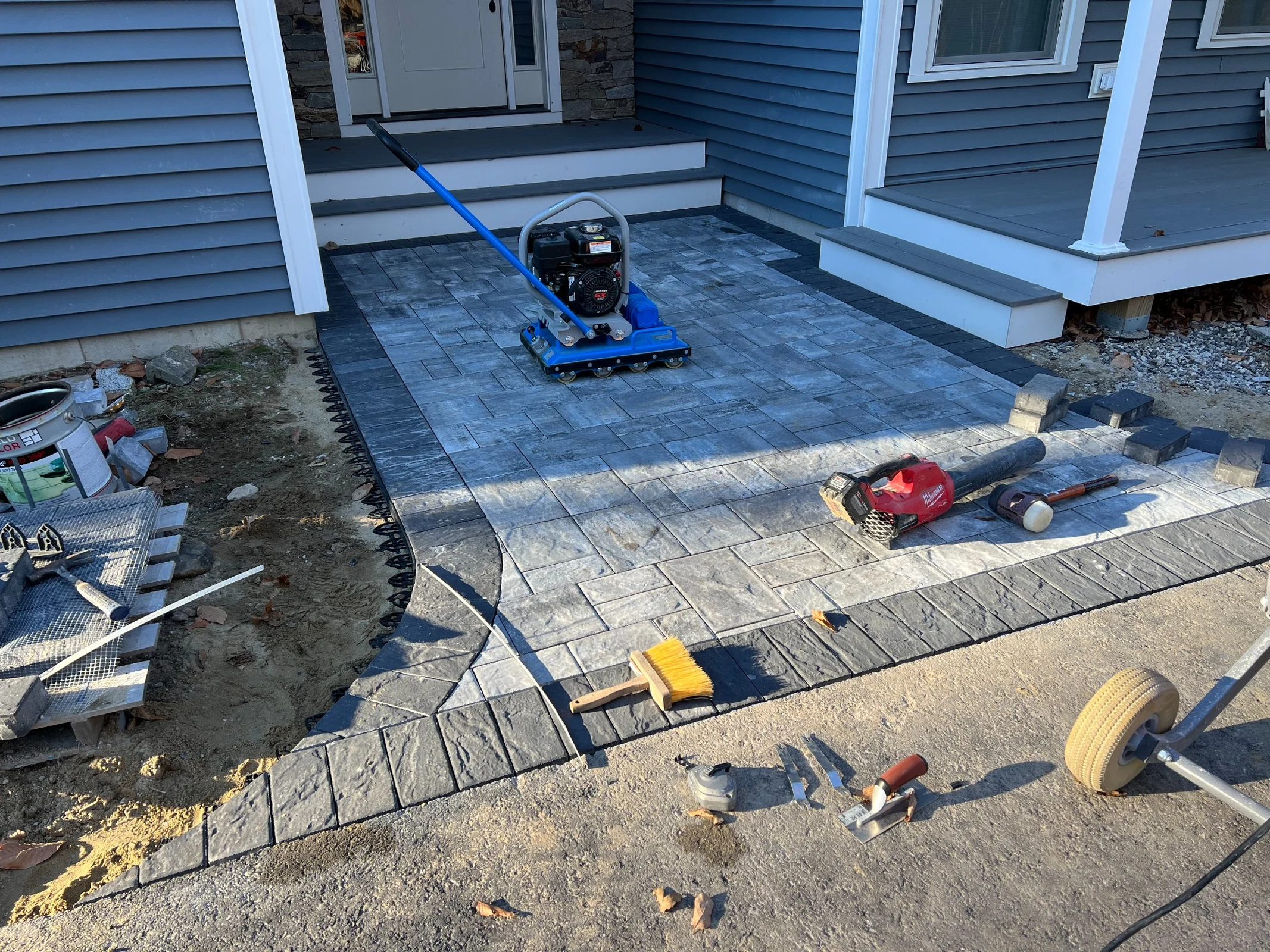Plate compactor setting a new gray paver patio with a charcoal border at the front steps of a home