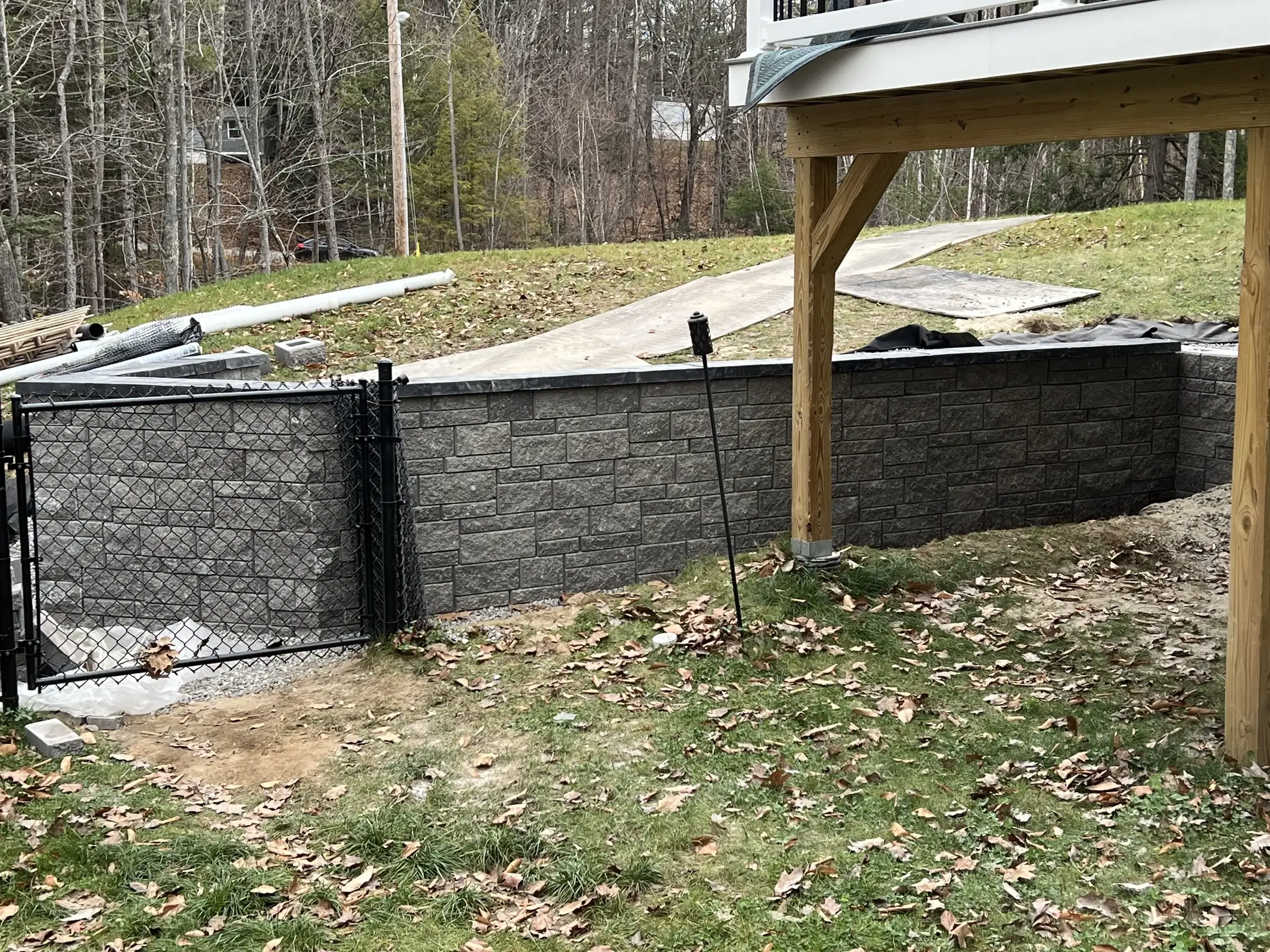 Charcoal segmental block retaining wall beneath a deck with a black chain-link fence and gate